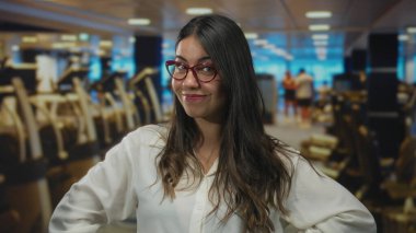 Spor salonu ortamında güvenle gülümseyen, arka planda bulanık egzersiz ekipmanları olan bir kadın, kapalı alanlarda gençlik enerjisi gösteriyor..