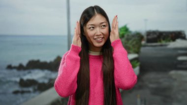 Deniz kenarındaki güvertede gülümseyen uzun saçlı pembe kazak giyen ve arka planda deniz manzaralı sakin ve neşeli bir anı yakalayan bir kadın..