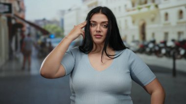 Uzun siyah saçlı bir kadın hareketli bir şehir caddesinde neşeli bir jest yapıyor, mavi bir gömlek giyiyor, şehir hayatının özünü yakalıyor..