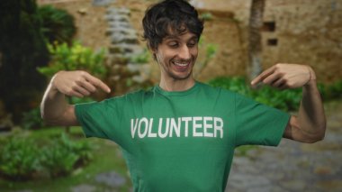 Adam gülümsüyor ve parkın dışındaki yeşil güneşli parkta gönüllü gömleği işaret ediyor..