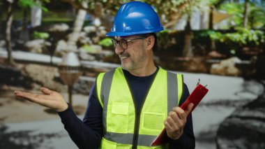 Güvenlik yeleği giymiş orta yaşlı bir adam açık şehir parkında elinde pano tutarken gülümsüyor..