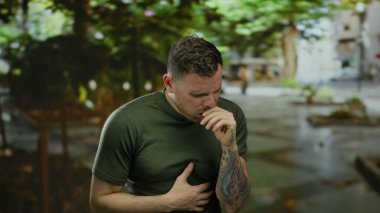 Şehir merkezinde hapşıran, dövmeleri görünen genç adam, bulanık sokak arka planının ortasında yeşil gömleğin altında rahatsızlığını ifade ediyor..