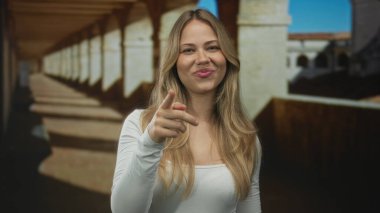 Sarışın kadın kamera karşısında üniversite manastırı koridorunu işaret ediyor..