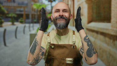 Sakallı ve eldiven takan bir adam, şehir sokaklarında neşeli bir ifadeyle iş önlüğü sergiliyor..