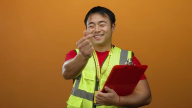 Yelekli genç adam turuncu arka plana kalp hareketi yapıyor..