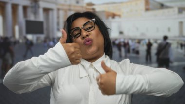 Vatikan şehir meydanında gülümseyen ve başparmak kaldıran kadın, çeşitli turistler ve tarihi mimari arkaplanının yer aldığı ikonik Avrupa ortamında neşeli bir an sergiliyor.