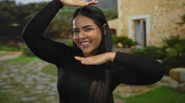 Elleriyle yüzünü çerçeveletmiş gülümseyen genç bir kadın eski bir kasabada kırsal taştan binalar ve yeşilliklerle dolu bir yerde duruyor..