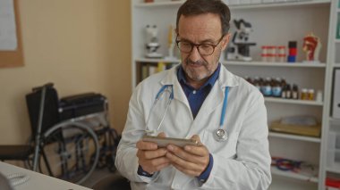 Klinikteki akıllı telefonu inceleyen doktor tıbbi ekipman ve tekerlekli sandalye ile çevrili laboratuvar önlüğü giyerek profesyonellik ve sağlık hizmetleri ortamını inceliyor..