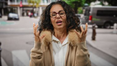 Kıvırcık saçlı bir kadın şehir sokaklarında gözlük ve açık havada kalın bir palto giyer..