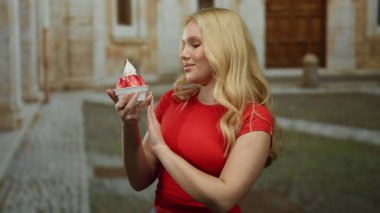 Sarışın genç bir kadın tarihi bir üniversite kampüsünde çilekli dondurma yiyor kırmızı bir gömlek giyiyor, güneşin altında sıcak bir şekilde gülümsüyor..