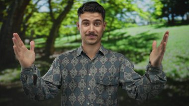 Güneşli bir günde, yeşil ağaçlar ve gölgelerle çevrili bir parkta el kol hareketi yapan genç bir İspanyol, desenli bir gömlek giymiş, elleri açık havada dışa dönük poz veriyor..