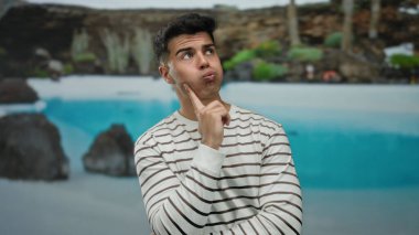 Çizgili gömlekli genç bir adam tatil köyü havuzunun yanında neşeli bir poz veriyor, otel ortamının boş havasını büyüleyici bir ifadeyle sergiliyor..