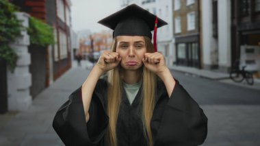 Mezuniyet elbisesi giymiş genç bir kadın alaycı bir şekilde şehir sokaklarında ağlıyor, mizah ve başarıyı şehir ortamında harmanlıyor..