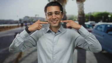 Genç adam bir şehir sokağında gülümseyip kendini işaret ediyor, gözlük ve saat takıyor, bulanık bir arka planda açık hava manzarası yaratıyor..