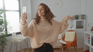 Bebek beşiği ve modern dekoru olan rahat bir yatak odasında, kazak ve kot pantolon giyen, akıllı telefon tutarken duygularını ifade eden bir kadın videosu..