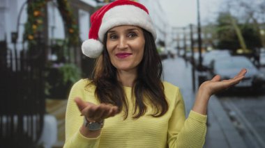 Noel Baba şapkalı kadın neşeli bir şekilde dışarıda, neşeli bir sokakta jest yapıyor, Noel sahnesine renkli bir dokunuş ekliyor..