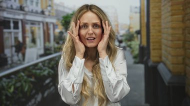 Beyaz tişörtlü, sarışın bir kadın sokakta poz veriyor. Şaşırdığını ya da şehir arka planında şok geçirdiğini söylüyor..