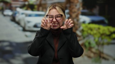 Gözlüklü, gülümseyen bir kadın park etmiş arabaları ve arka planda yeşilliği olan bir şehir caddesinde neşeli pençeler yapıyor..