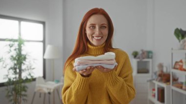 Kızıl saçlı kadın katlanmış giysiler tutuyor. Parlak bir çamaşır odasında gülümsüyor. Sıcak bir ev atmosferi var..