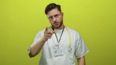 Sakallı, beyaz, genç bir doktor. Parmak ucunda, izole edilmiş sarı arka plana karşı steteskop takıyor. Ciddi bir ifade ve profesyonel bir varlık sergiliyor..