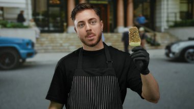 Önlüklü, sakallı genç garson. Caddede dondurma tutuyor. Gülümsüyor ve açık havada eldiven takıyor..