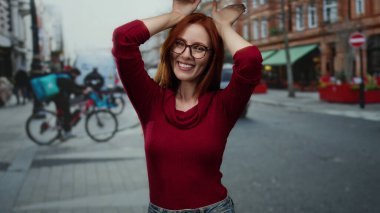 Kızıl saçlı bir kadın gözlüklü ve kırmızı kazaklı bir kadın, şehir aktiviteleriyle çevrili, neşeli bir yetişkin cazibesi yayıyor..