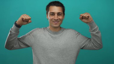 Neşeli ve kendine güvenen genç bir adam canlı deniz mavisi arkaplanına karşı yumruklarını sıkarak başarı ve coşkuyla iletişim kuruyor..