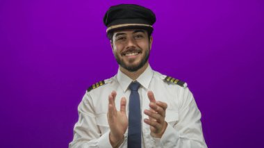 Üniformalı genç adam canlı mor bir arka plana karşı alkışlıyor, kendine güvenen, sakallı ve şapkalı, profesyonel ve enerjik bir tavır sergiliyor..