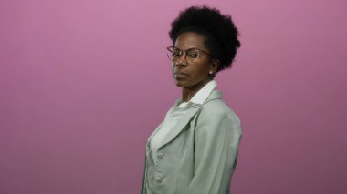 İzole edilmiş pembe arka plana karşı duran, beyaz gömlek üzerine gözlük ve ceket giyen bir kadın.