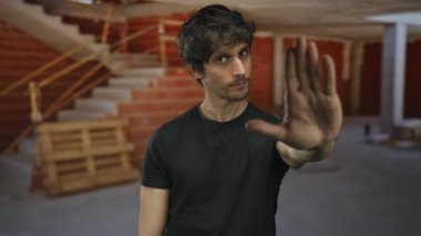 Delikanlı, tamamlanmamış binada tuğla ve ahşap paletin ortasında durup el hareketi yapıyor; meydan okuma uyarısı.