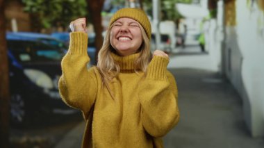 Sarı süveter ve bereyle gülen kadın şehir merkezinde gündüz vakti mutluluğunu ve neşesini ifade ediyor..