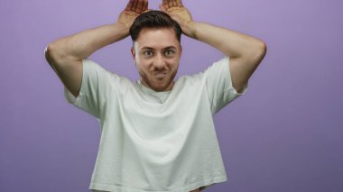 Sakallı genç bir adam, mor bir arka planda, elleri başının üstünde, eğlenceli, kaprisli bir ifade ve neşe sahnesi yaratıyor.