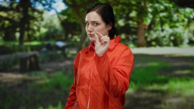 Turuncu hapisteki kadın dışarıda yeşil ve güneş ışığıyla çevrili bir parkta özgürlük ve hapsedilme arasında bir zıtlık hissi yayıyor..