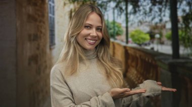 Sarı saçlı ve gülümseyen yüzlü bir kadın avuçlarını dar bir sokakta eski bir duvarın ve sağlam bir korkuluğun yanında sunuyor..