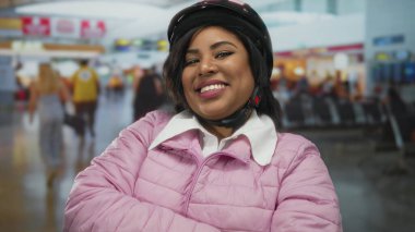 Pembe ceketli, kasklı, havaalanı terminalinde gülümseyen bir kadın güven ve pozitiflik gösteriyor..