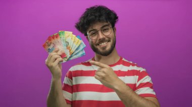 Çizgili tişörtlü, sakallı, parlak pembe arkaplanlı İsviçre frangına gülümseyen ve işaret eden genç İspanyol adam coşku ve gururla renkli para birimi notları gösteriyor..