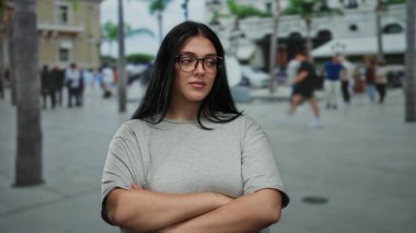 Genç bir kadın kentin işlek bir caddesinde kendine güvenerek duruyor, canlı bir şehir zeminine karşı geniş beden ve şık gözlüklerini sergiliyor..