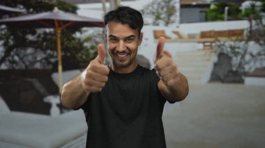 Genç, İspanyol asıllı, neşeli bir gülümsemeyle baş parmak kaldıran, yeşil ve beyaz duvarlı, güneşli havayla çevrili, yumuşak odaklı bir avlu..