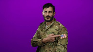 Askeri üniformalı İspanyol sakallı adam parlak bir mor arka plana karşı Amerikan bayrak bandını işaret ediyor..