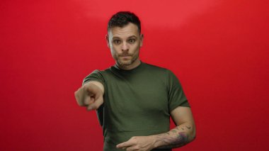 Yeşil tişörtlü genç İspanyol adam canlı kırmızı bir arka plana karşı ciddi bir ifadeyle işaret ediyor, yakışıklı ve çekici özelliklerini izole bir ortamda sergiliyor..
