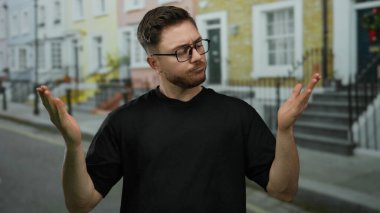 Sakallı ve gözlüklü genç bir adam şehir sokaklarında, açık avuçlarla kafa karışıklığını dile getiriyor..