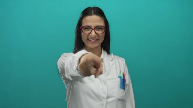 Laboratuvar önlüklü İspanyol kadın bilim adamı, yeşil bir zemine el hareketi yapıyor, gülümsüyor ve gözlük takıyor, pozitifliğini ve profesyonelliğini ifade ediyor..