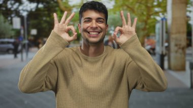 Erkek, açık havada güneş ışığında iyi bir sembol oluşturmak için ellerini kaldırır. İspanyol adam, açık bir şekilde onaylamak için sevinçle gülümsüyor..