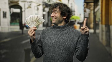 Hispanik bir adam elinde Rus banknotları tutuyor ve canlı bir şehir caddesinde gökyüzünü işaret ediyor. Açık hava ortamında akıllıca bir finansal karardan emin bir şekilde gülümsüyor..