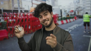 İspanyol genç adam güneşli bir günde elinde kronometreyle yol inşaatının arka planında gülümseyerek zamanı ve şehir enerjisini vurguluyor..
