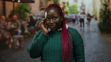 Kırmızı örgülü yeşil kazaklı kadın şehir caddesindeki terasta duruyor. Etrafı hareketli şehir restoranları ve kafelerle çevrili. Şehir yaşamını somutlaştırıyor..