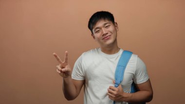 Mavi sırt çantalı genç Çinli adam gülümsüyor ve izole edilmiş kahverengi arka plan duvarına barış işareti yapıyor..