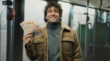 Elinde Türk lira banknotları tutan İspanyol bir adam otobüste gülümsüyor, kapalı alanda bir finansal başarı ve seyahat sahnesini vurguluyor..
