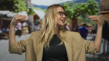 Kadın şehir caddesinde konuşurken, bej bir ceket giyiyor, açık havada canlı bir şekilde yayalar tarafından çevrelenmiş..