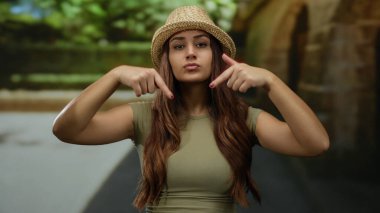 Güneşli bir caddeyi işaret eden şapkalı genç bir kadın. Arka planda yeşil ve taş binalar var..